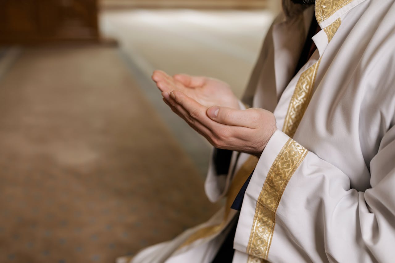 Home A man in traditional Middle Eastern attire prays indoors, hands clasped together.
